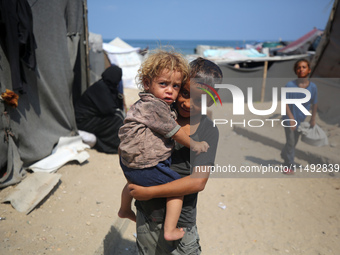 A displaced Palestinian boy is carrying his younger brother while standing next to their tent near the beach in Deir al-Balah in the central...