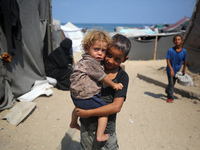 A displaced Palestinian boy is carrying his younger brother while standing next to their tent near the beach in Deir al-Balah in the central...