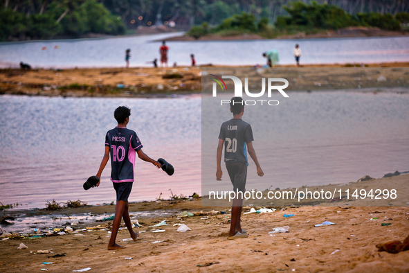 Boys are walking towards a football game as youth are playing a game of football (soccer) by the ocean at sunset along Paruthiyoor Beach in...