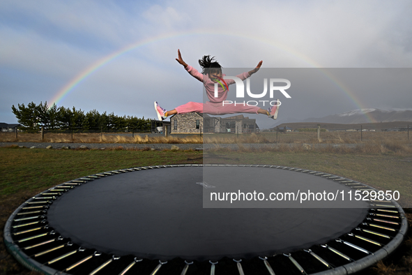 A little girl, Minudi Menulya, jumps on a trampoline as a rainbow glows in the background in Twizel, New Zealand, on August 31, 2024. 