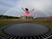 A little girl, Minudi Menulya, jumps on a trampoline as a rainbow glows in the background in Twizel, New Zealand, on August 31, 2024. (