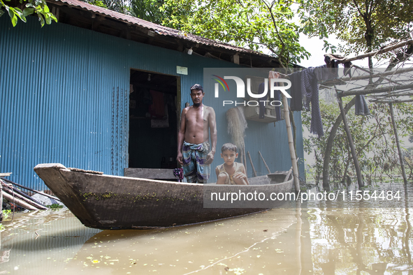 A man stands in front of his flooded house, and his young child sits on a boat, the only means of transport in a village in Choumuhani Munic...