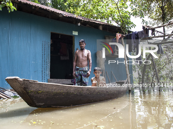 A man stands in front of his flooded house, and his young child sits on a boat, the only means of transport in a village in Choumuhani Munic...