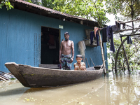 A man stands in front of his flooded house, and his young child sits on a boat, the only means of transport in a village in Choumuhani Munic...