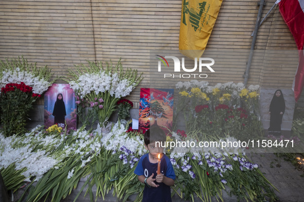 A young Iranian boy holds a candle while standing next to portraits of a young Lebanese girl who is killed in a deadly pager attack, a flag...