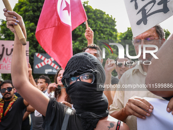 A young female demonstrator with her face covered by a bandanna raises her arm as others wave the national flag of Tunisia during a demonstr...