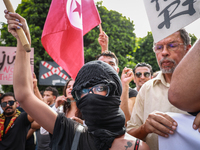 A young female demonstrator with her face covered by a bandanna raises her arm as others wave the national flag of Tunisia during a demonstr...