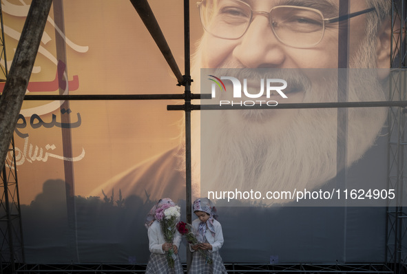 Two young Iranian girls hold flowers while standing together under a giant banner featuring a portrait of Lebanon's Hezbollah Secretary Gene...