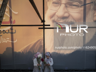 Two young Iranian girls hold flowers while standing together under a giant banner featuring a portrait of Lebanon's Hezbollah Secretary Gene...