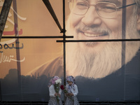 Two young Iranian girls hold flowers while standing together under a giant banner featuring a portrait of Lebanon's Hezbollah Secretary Gene...