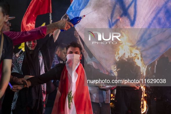 An Iranian man wears a Lebanese flag while protesters burn an Israeli flag during a celebration for Iran's missile attack against Israel in...