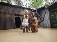 An elderly husband and wife stand in front of their house in knee-deep flood water in a village in Jhenaigati upazila of Sherpur district in...