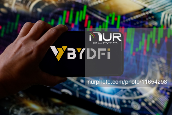 In this photo illustration, the BYDFI logo is seen displayed on a smartphone screen against a computer screen displaying a stock market grap...