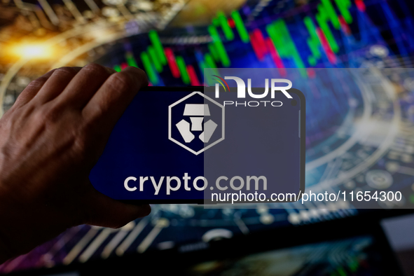 In this photo illustration, the Crypto.com logo is seen displayed on a smartphone screen against a computer screen displaying a stock market...