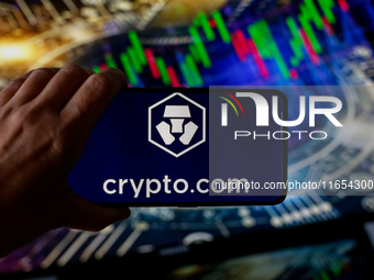 In this photo illustration, the Crypto.com logo is seen displayed on a smartphone screen against a computer screen displaying a stock market...