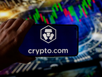 In this photo illustration, the Crypto.com logo is seen displayed on a smartphone screen against a computer screen displaying a stock market...