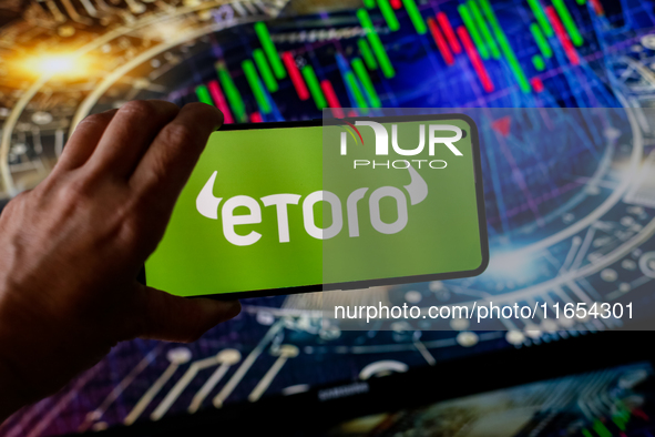 In this photo illustration, the eToro logo is seen displayed on a smartphone screen against a computer screen displaying a stock market grap...