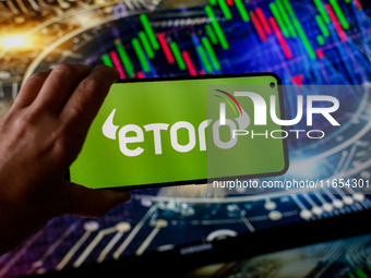 In this photo illustration, the eToro logo is seen displayed on a smartphone screen against a computer screen displaying a stock market grap...