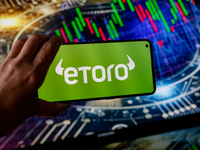 In this photo illustration, the eToro logo is seen displayed on a smartphone screen against a computer screen displaying a stock market grap...