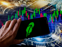 In this photo illustration, the Robinhood logo is seen displayed on a smartphone screen against a computer screen displaying a stock market...