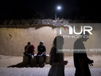 Iranian youths walk past young men sitting on a path in Shiraz, Iran, on October 7, 2024, at night.  (