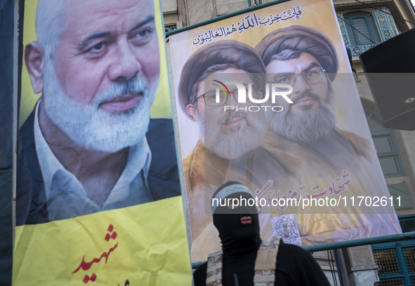 An Islamic Revolutionary Guard Corps (IRGC) personnel, wearing a mask and a uniform symbolizing a Hamas warrior, stands under portraits of L...