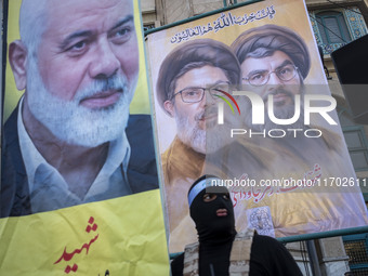 An Islamic Revolutionary Guard Corps (IRGC) personnel, wearing a mask and a uniform symbolizing a Hamas warrior, stands under portraits of L...