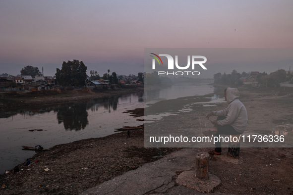 A man sits on a concrete block and watches Kashmiri boys catch fish on a cold evening in River Jhelum in Sopore, Jammu and Kashmir, India, o...