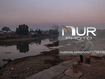 A man sits on a concrete block and watches Kashmiri boys catch fish on a cold evening in River Jhelum in Sopore, Jammu and Kashmir, India, o...