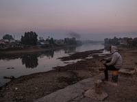 A man sits on a concrete block and watches Kashmiri boys catch fish on a cold evening in River Jhelum in Sopore, Jammu and Kashmir, India, o...