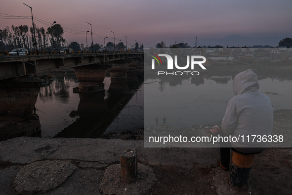 A man sits on a concrete block and watches Kashmiri boys catch fish on a cold evening in River Jhelum in Sopore, Jammu and Kashmir, India, o...