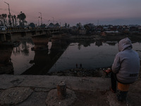 A man sits on a concrete block and watches Kashmiri boys catch fish on a cold evening in River Jhelum in Sopore, Jammu and Kashmir, India, o...