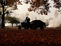 A person wears a jacket as he rides a tractor while women burn chirnar leaves to make charcoal during the autumn season in Sopore, Jammu and...