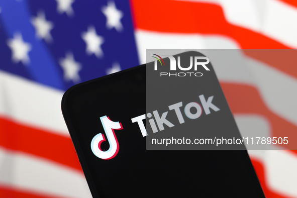 The logo of the social network TikTok appears on the screen of a smartphone, and in the background, the flag of the United States is display...