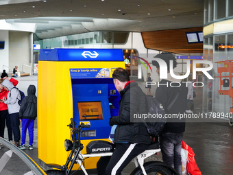 Two men interact with a bright yellow and blue ticket machine from the Dutch Railways (NS) inside Arnhem Central Station in Arnhem, Netherla...