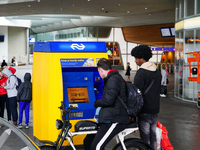 Two men interact with a bright yellow and blue ticket machine from the Dutch Railways (NS) inside Arnhem Central Station in Arnhem, Netherla...