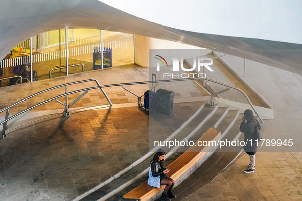 A traveler sits on a curved wooden bench, engrossed in her phone, while another stands nearby, seemingly in conversation, at Arnhem Centraal...