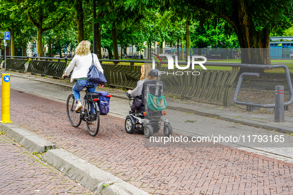 A bicyclist and a person in a motorized wheelchair share a smooth, brick-paved bike path bordered by lush trees and urban facilities in Venl...