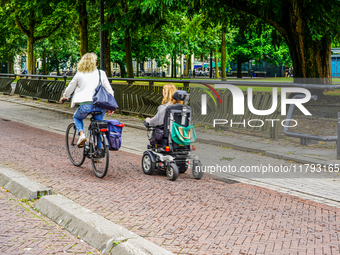 A bicyclist and a person in a motorized wheelchair share a smooth, brick-paved bike path bordered by lush trees and urban facilities in Venl...