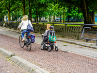 A bicyclist and a person in a motorized wheelchair share a smooth, brick-paved bike path bordered by lush trees and urban facilities in Venl...