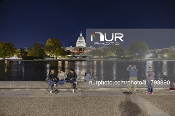 People taking photos and relaxing in front of the water. Night view of the illuminated US Capitol, the seat of the United States Congress wi...