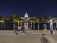 People taking photos and relaxing in front of the water. Night view of the illuminated US Capitol, the seat of the United States Congress wi...