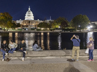 People taking photos and relaxing in front of the water. Night view of the illuminated US Capitol, the seat of the United States Congress wi...