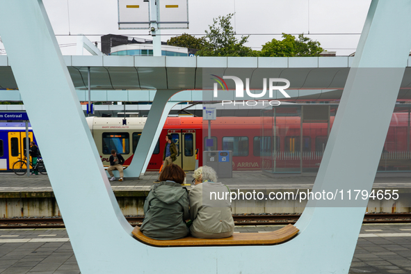 In Arnhem, Netherlands, on July 27, 2023, two passengers sit on a modern bench at Arnhem Central Station. In the background, an NS Sprinter...