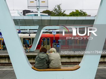 In Arnhem, Netherlands, on July 27, 2023, two passengers sit on a modern bench at Arnhem Central Station. In the background, an NS Sprinter...