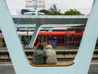 In Arnhem, Netherlands, on July 27, 2023, two passengers sit on a modern bench at Arnhem Central Station. In the background, an NS Sprinter...