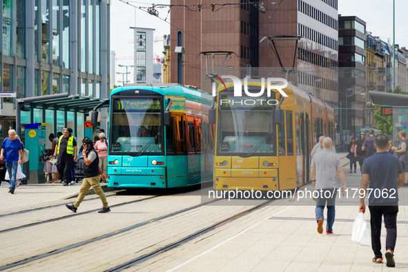 At a tram stop in a city, people board and alight two brightly colored trams, turquoise and yellow, on August 15, 2023. The platform is aliv...