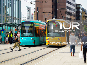 At a tram stop in a city, people board and alight two brightly colored trams, turquoise and yellow, on August 15, 2023. The platform is aliv...