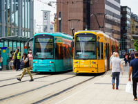 At a tram stop in a city, people board and alight two brightly colored trams, turquoise and yellow, on August 15, 2023. The platform is aliv...