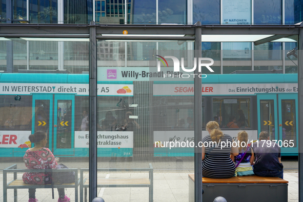 Passengers sit on benches at a tram stop near Willy-Brandt-Platz in a modern city setting on August 15, 2023. Through the glass panels, a tu...